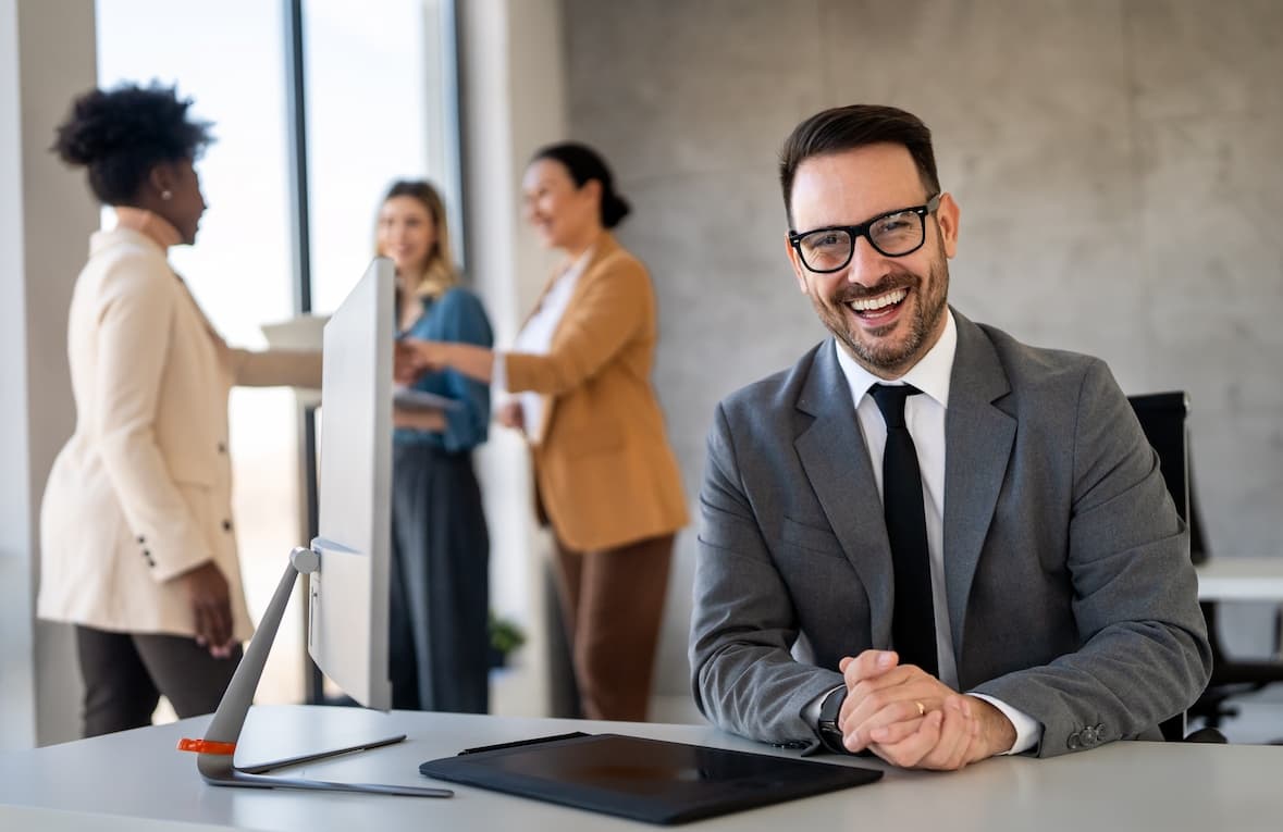 A smiling professional at his desk with colleagues in the background