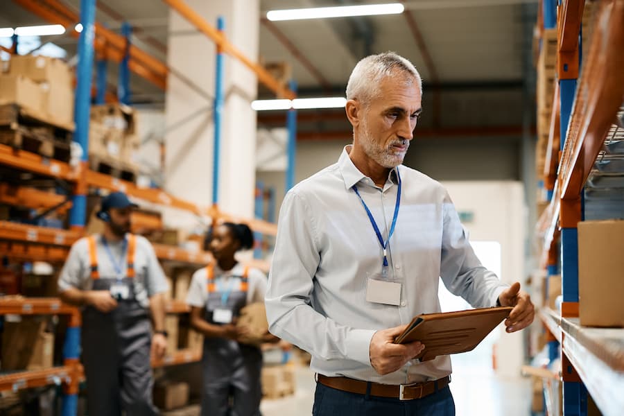 Mature warehouse manager using digital tablet at storage compartment.