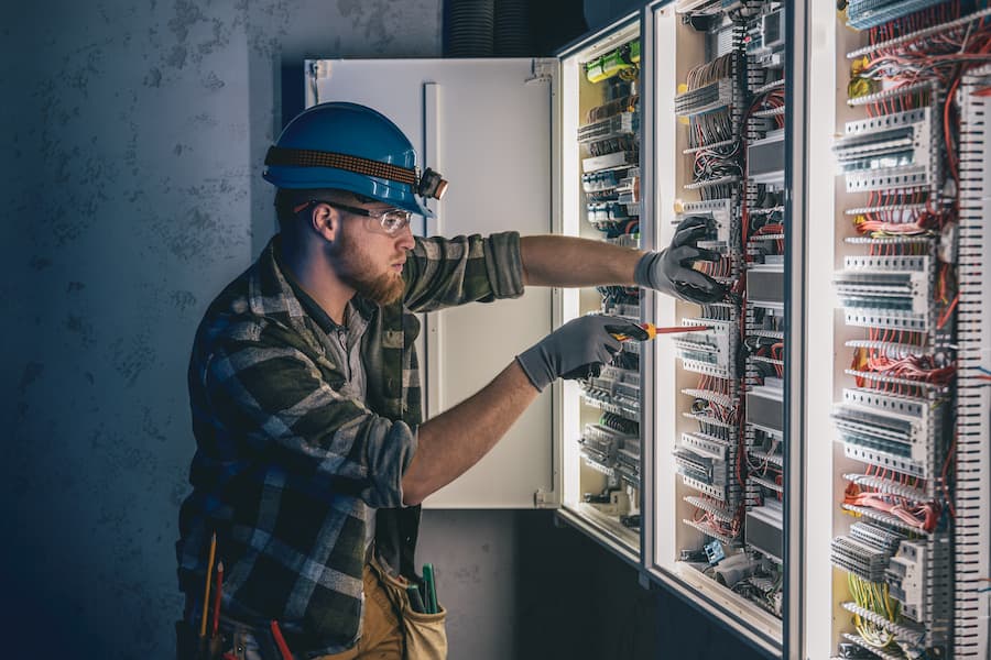 A male electrician works in a switchboard in overalls against the backdrop of emergency lighting.
