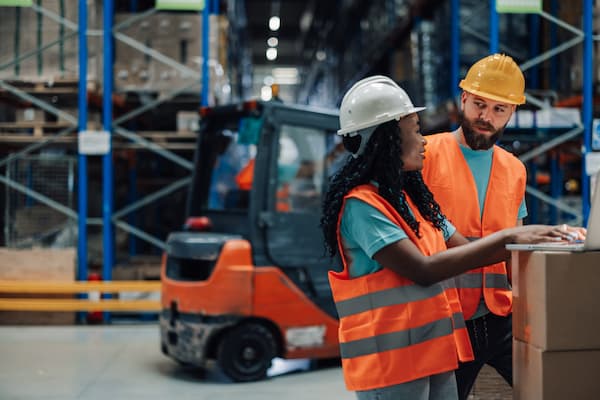 Warehouse workers using laptop next to forklift are discussing logistics